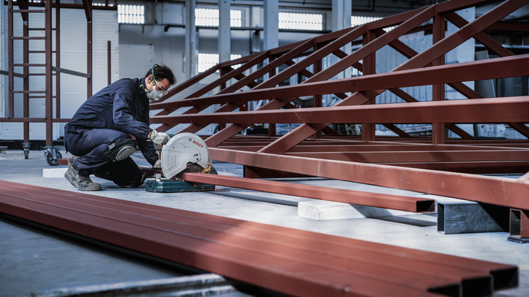 안전 장비를 착용한 사람이 공장에서 원형 톱을 이용해 강철 보를 자르고 있습니다.