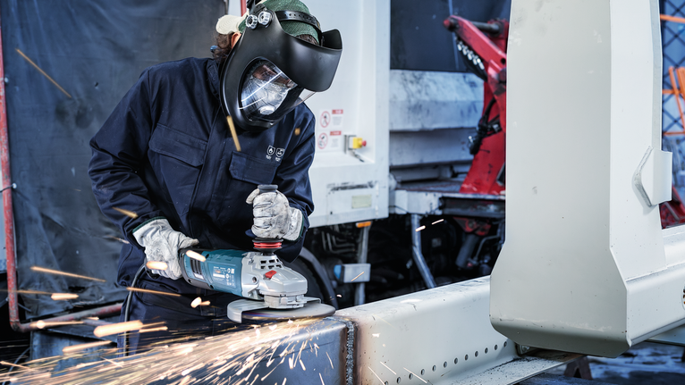 안전 장비를 착용한 사람이 각도 분쇄기를 사용하여 금속 빔을 자릅니다.