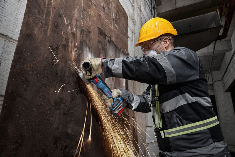 안전 장비를 착용한 사람이 무선 각도 그라인더로 녹슨 금속판을 갈고 있습니다.