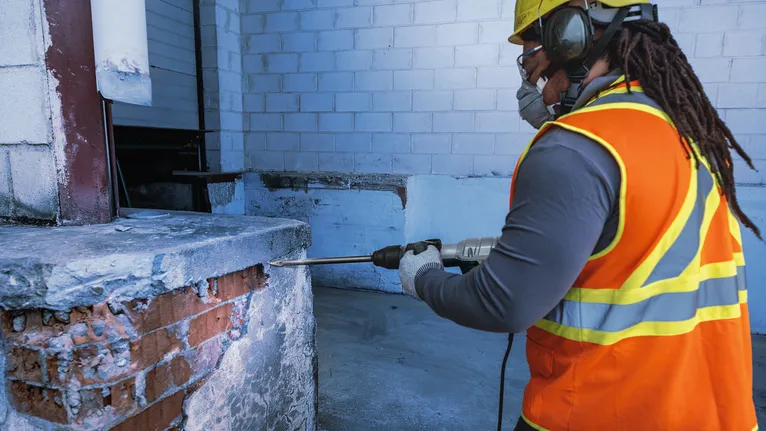 안전장비를 착용한 사람이 건설 현장에서 콘크리트 벽에 드릴을 박고 있다.