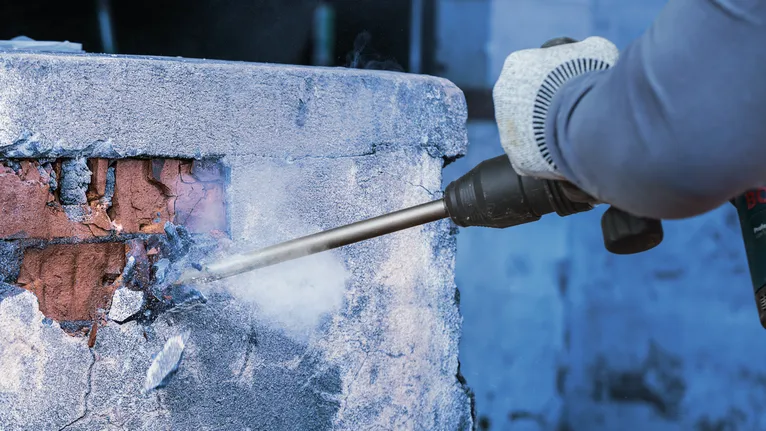 안전 장비를 착용한 사람이 철거 도구를 사용하여 콘크리트 벽을 부수고 있습니다.