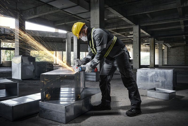 안전 장비를 착용한 사람이 건설 현장에서 금속 덕트에 각도 그라인더를 사용하고 있습니다.