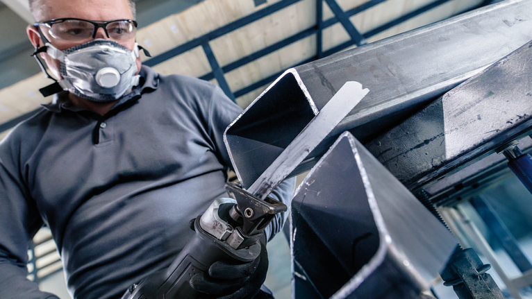 안전 장비를 착용한 사람이 왕복톱을 사용해 금속 막대를 자릅니다.