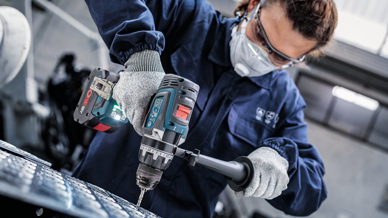 안전 장비를 착용한 사람이 무선 전동 공구로 금속에 구멍을 뚫고 있습니다.