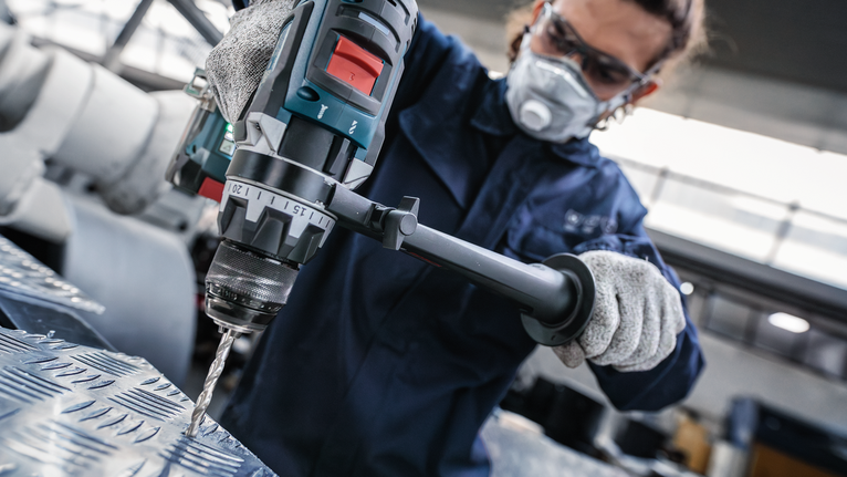 안전 장비를 착용한 사람이 전동 공구를 사용하여 질감이 있는 금속에 구멍을 뚫고 있습니다.