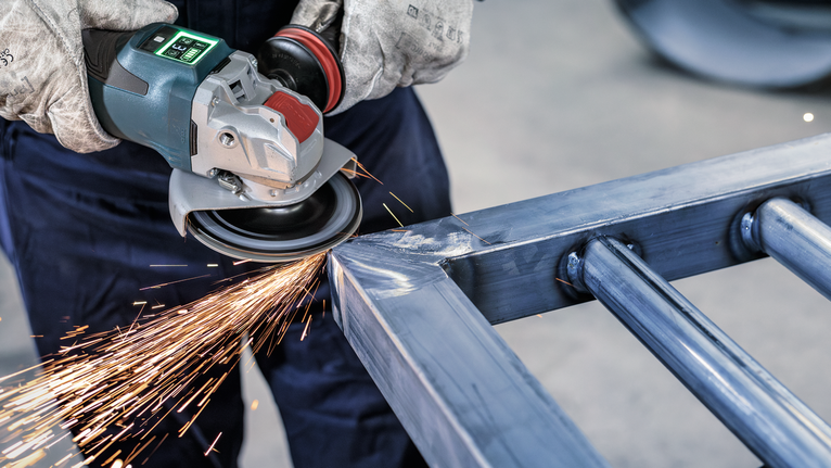 안전 장비를 착용한 사람이 각도 그라인더로 금속 프레임을 갈아서 불꽃을 낸다.