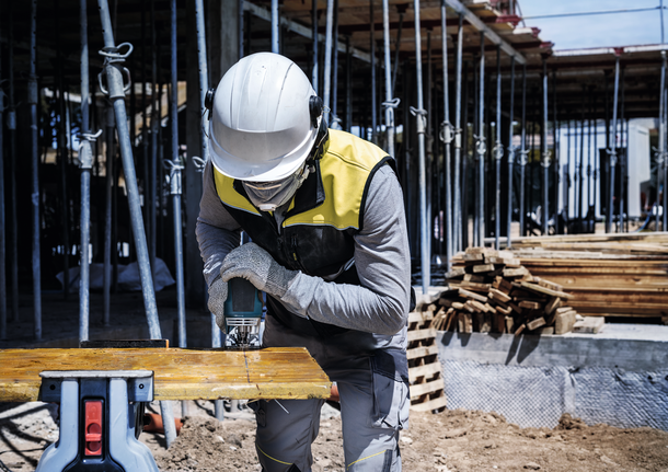 안전 장비를 착용한 사람이 건설 현장에서 전동 공구를 이용해 나무 판자를 자르고 있습니다.