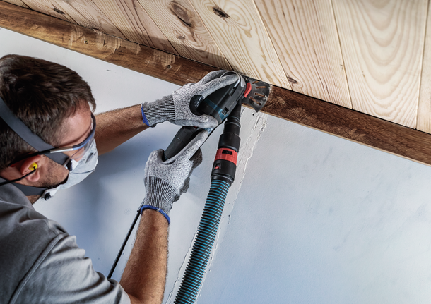 안전장비를 착용한 사람이 전동공구를 이용해 나무 천장 모서리를 샌딩하고 있습니다.