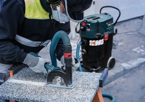 안전 장비를 착용한 사람이 먼지 추출기에 연결된 원형 톱을 이용해 돌을 자릅니다.