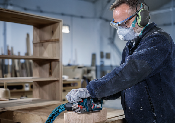 안전 장비를 착용한 사람이 작업장에서 나무판을 사포질하고 있습니다.