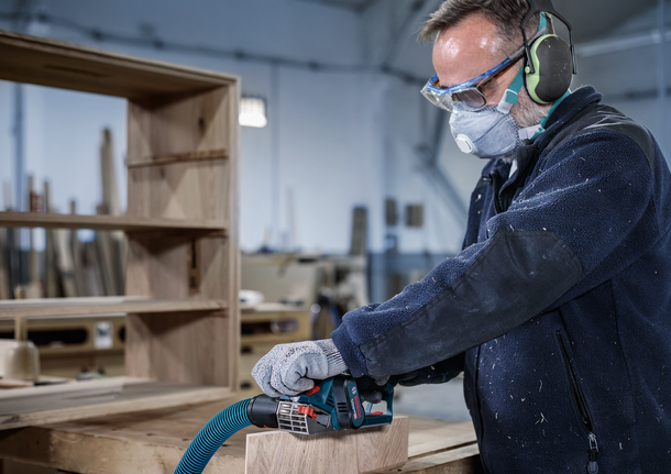 안전 장비를 착용한 사람이 작업장에서 나무판을 사포질하고 있습니다.