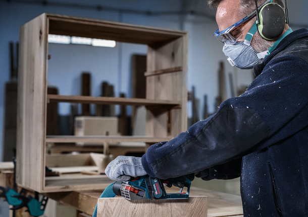 안전 장비를 착용한 사람이 전동 공구로 나무 가구를 연마하고 있습니다.
