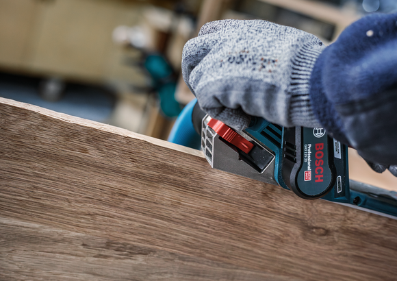 안전 장비를 착용한 사람이 전동 공구를 이용해 나무판의 가장자리를 매끄럽게 다듬고 있습니다.