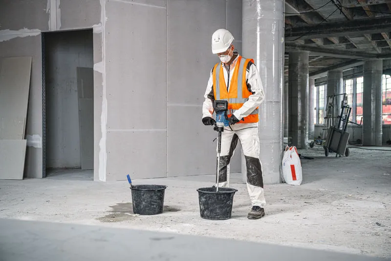 안전 장비를 착용한 사람이 교반 장치를 사용해 양동이에 있는 재료를 섞습니다.