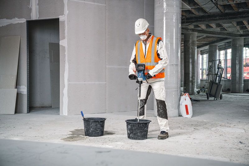 안전 장비를 착용한 사람이 교반 장치를 사용해 양동이에 있는 재료를 섞습니다.