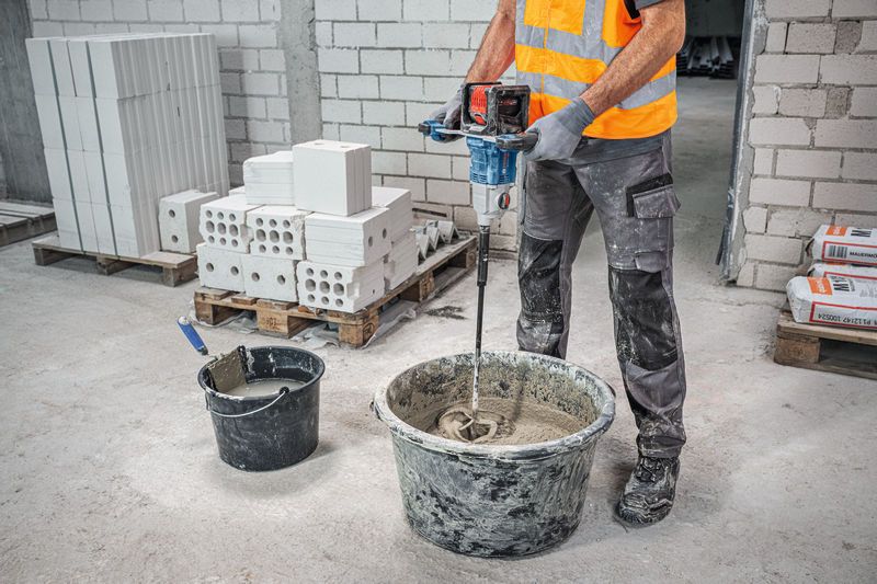 안전 장비를 착용한 사람이 교반 장치가 있는 양동이에 모르타르를 섞고 있습니다.