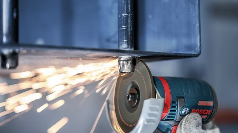 안전 장비를 착용한 장갑낀 작업자가 각도 분쇄기로 금속 빔을 갈아서 불꽃을 낸다.