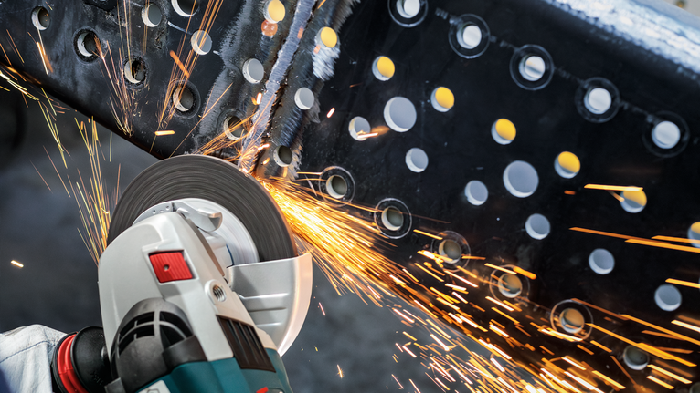 안전 장비를 착용한 사람이 전동 공구로 금속 막대를 갈아서 불꽃을 낸다.