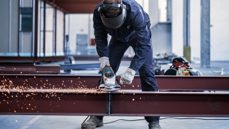 안전 장비를 착용한 사람이 강철 빔을 갈아서 밝은 불꽃을 낸다.