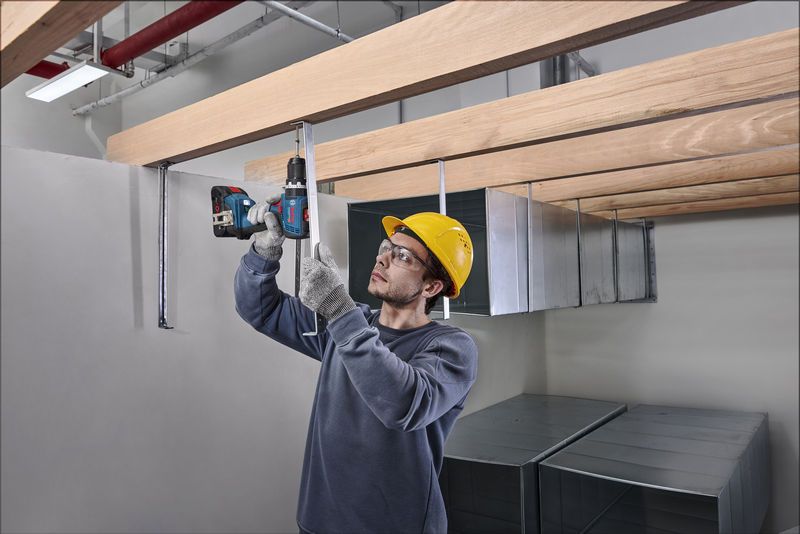 안전장비를 착용한 사람이 나무 들보 아래에 있는 금속 막대에 구멍을 뚫고 있습니다.