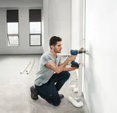 한 사람이 밝고 미완성된 방에 파이프를 설치하기 위해 벽에 구멍을 뚫고 있습니다.