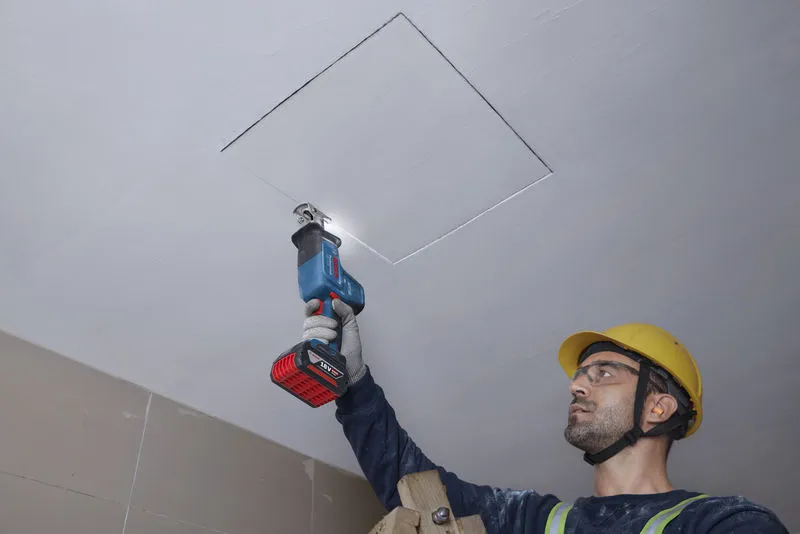 안전 장비를 착용한 사람이 왕복톱으로 천장 패널을 자릅니다.