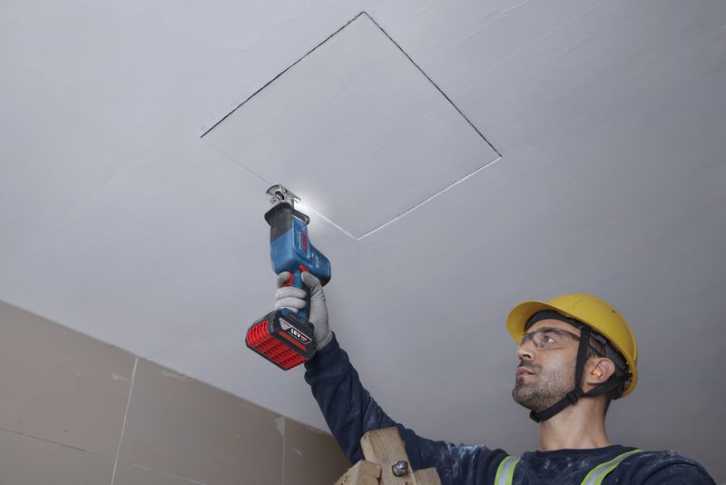 안전 장비를 착용한 사람이 왕복톱으로 천장 패널을 자릅니다.