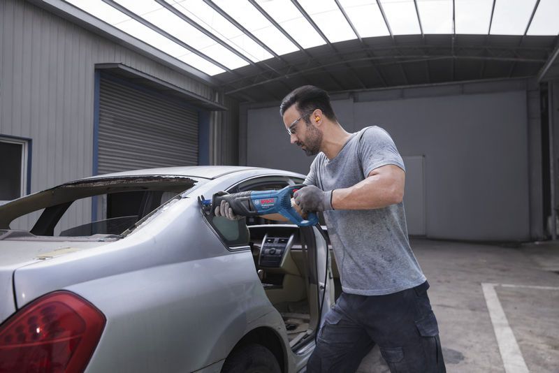 안전 장비를 착용한 사람이 왕복톱을 사용하여 자동차의 뒷창틀을 자르고 있습니다.