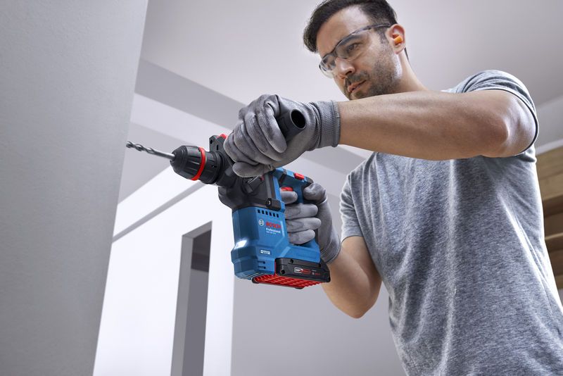 안전장비를 착용한 사람이 무선 회전 망치를 이용해 콘크리트 벽에 구멍을 뚫고 있습니다.