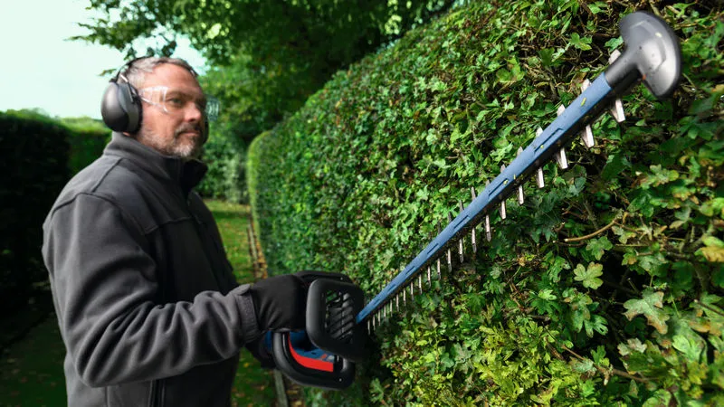 GHE 18V-50 (II) 안전장비를 착용한 사람이 무선 울타리 트리머로 울타리를 다듬고 있습니다.