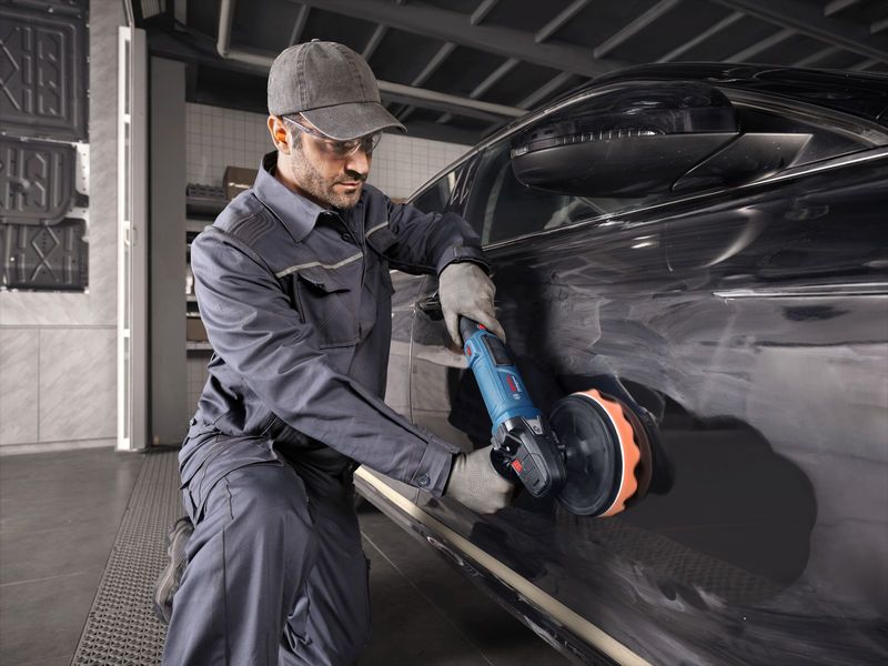 안전장비를 착용한 사람이 전동공구를 이용해 검은색 자동차를 닦고 있다.