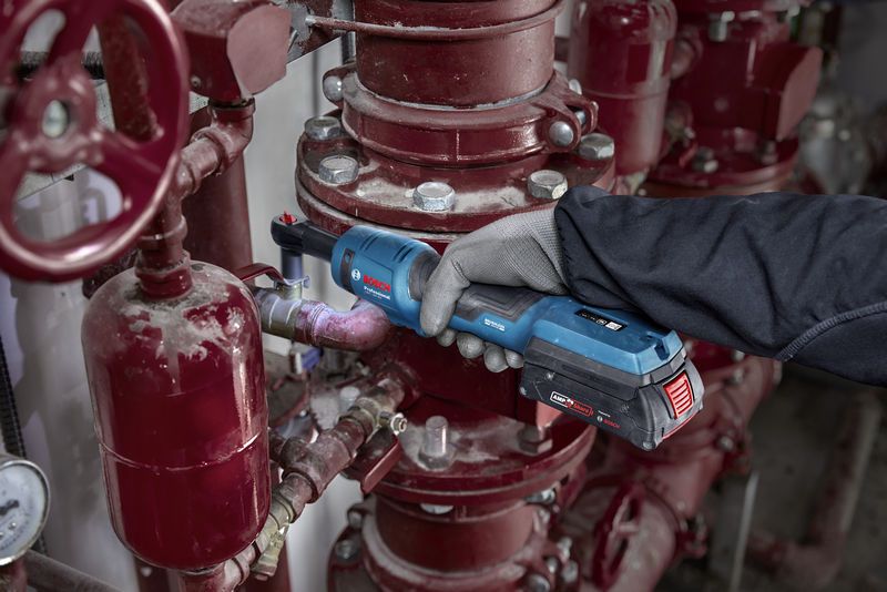 안전 장비를 착용한 사람이 산업용 배관에 무선 래칫 렌치를 사용하고 있습니다.