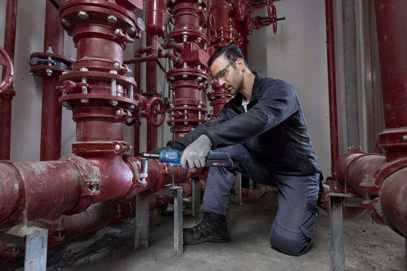 안전 장비를 착용한 사람이 빨간색 파이프 시스템에서 무선 래칫 렌치를 사용하고 있습니다.