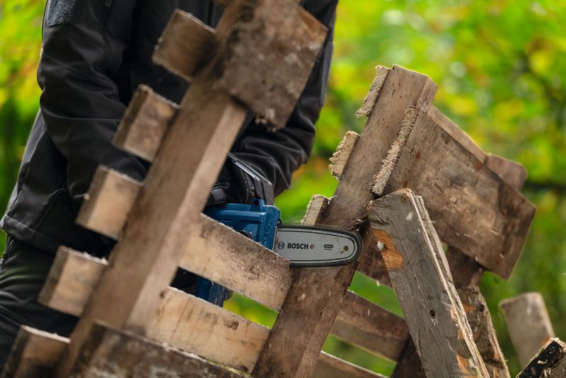 안전장비를 착용한 사람이 무선 가지치기 톱을 사용하여 나무 판자를 자르고 있습니다.