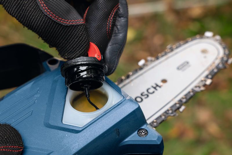 안전 장비를 착용한 사람이 무선 가지치기 톱의 칼날 근처에 있는 오일 캡을 엽니다.