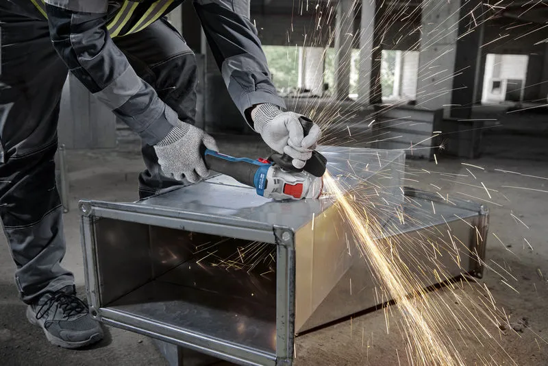 안전 장비를 착용한 사람이 각도 분쇄기로 금속 덕트를 갈아서 불꽃을 발생시킵니다.