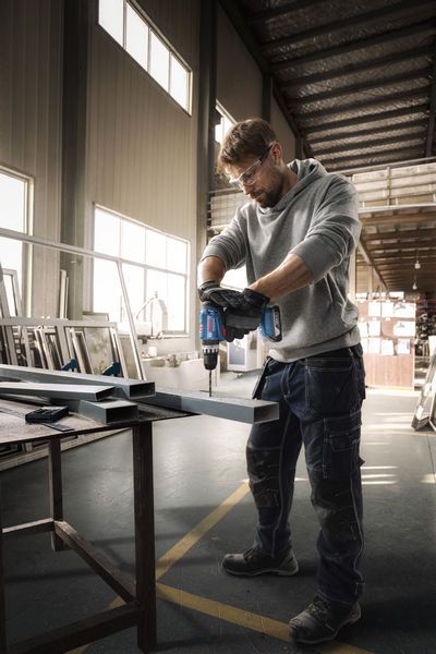 안전 장비를 착용한 사람이 작업장에서 금속 보에 구멍을 뚫고 있다.