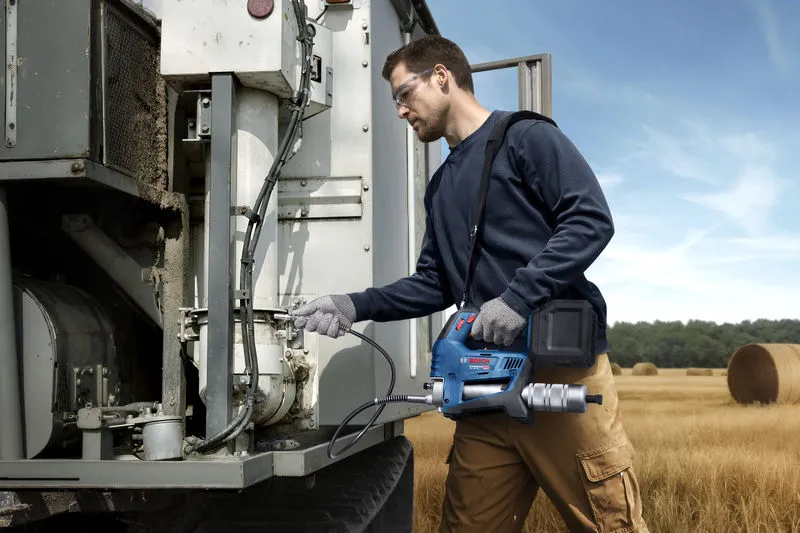 안전 장비를 착용한 사람이 무선 그리스 건을 사용해 농기계를 정비하고 있습니다.