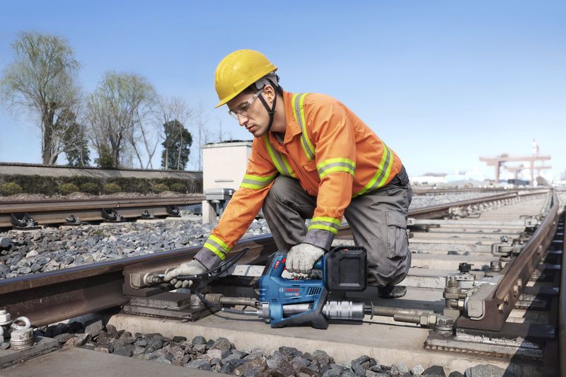 안전장비를 착용한 사람이 무선 그리스 건을 사용해 철도 선로를 유지관리하고 있습니다.