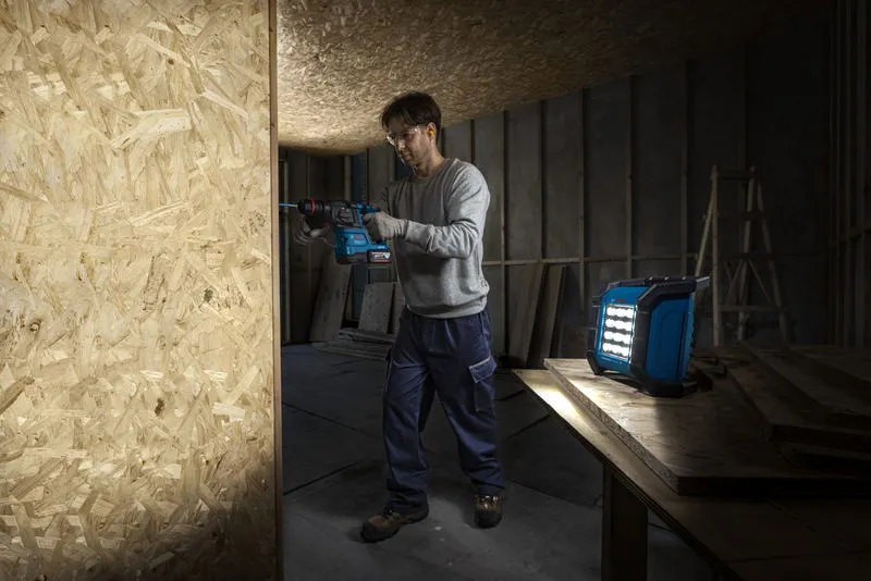 안전 장비를 착용한 사람이 무선 작업 현장 조명 근처의 나무판에 구멍을 뚫고 있습니다.