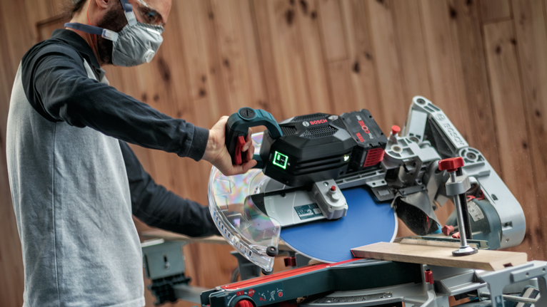 안전 장비를 착용한 사람이 마이터 톱을 작동하여 나무판을 자르고 있습니다.