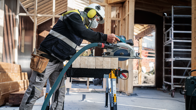 안전 장비를 착용한 사람이 건설 현장에서 원형 톱으로 목재 들보를 자르고 있습니다.