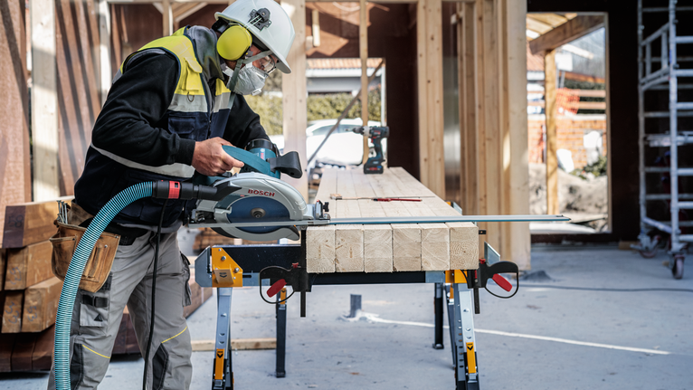 안전 장비를 착용한 사람이 건설 현장에서 원형 톱을 사용하여 나무 들보를 자르고 있습니다.