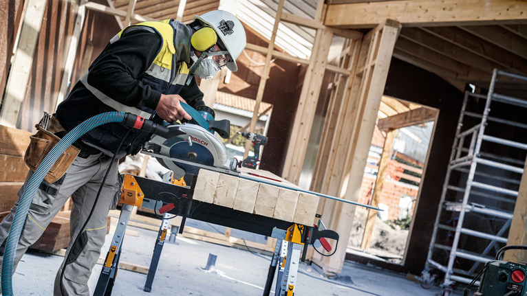 안전 장비를 착용한 사람이 건설 현장에서 원형 톱을 사용하여 나무 판자를 자르고 있습니다.