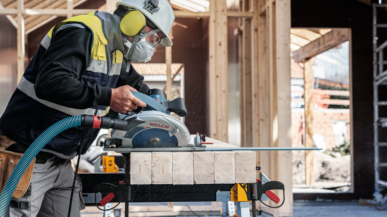 안전 장비를 착용한 사람이 건설 현장에서 원형 톱으로 목재를 자르고 있습니다.