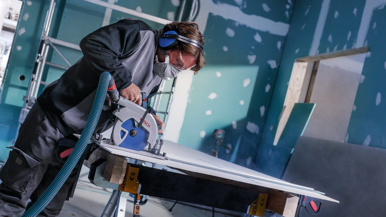 안전 장비를 착용한 사람이 건설 현장에서 원형 톱으로 판자를 자르고 있습니다.