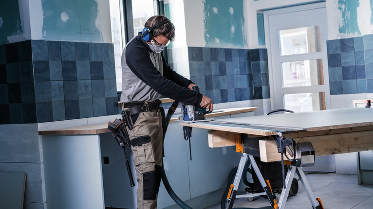 안전 장비를 착용한 사람이 타일이 깔린 방에서 전동 공구를 사용해 판자를 자르고 있습니다.