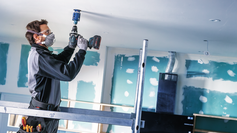 안전 장비를 착용한 사람이 건설 현장에서 천장에 구멍을 뚫고 있습니다.