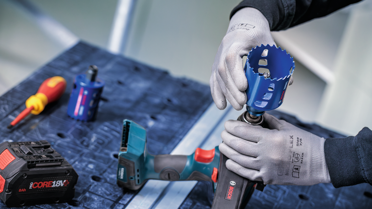 안전 장비를 착용한 사람이 작업대 위에 있는 무선 드릴에 홀 톱을 부착하고 있습니다.