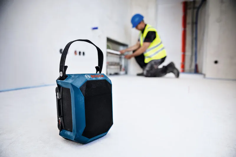 건설 현장 바닥에 블루투스 스피커가 있고, 배경에는 안전 장비를 착용한 사람이 있습니다.